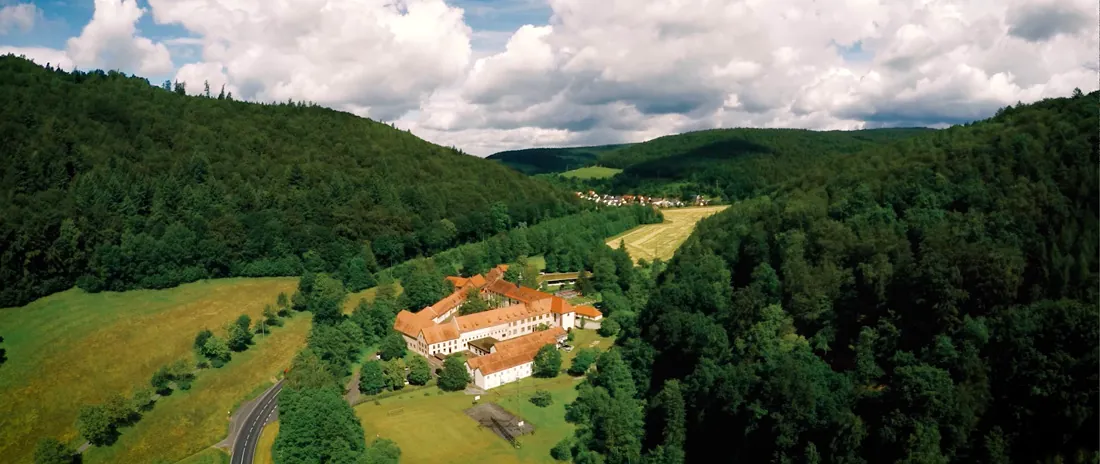 Luftaufnahme: die Klosteranlage mit roten Dächern zwischen bewaldeten Hängen im Elsavatal